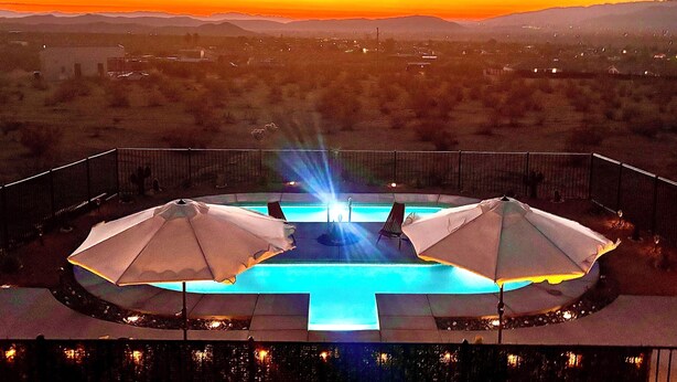 A pool with two umbrellas, surrounded by a fence, overlooking a desert landscape with mountains in the distance. - Magical Vibes on 5 Acres with panoramic views. (Joshua Tree)