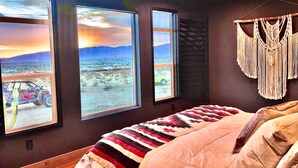 A bedroom with a large bed, a macrame wall hanging, and three windows offering a view of a desert landscape. - Magical Vibes on 5 Acres with panoramic views. (Joshua Tree)