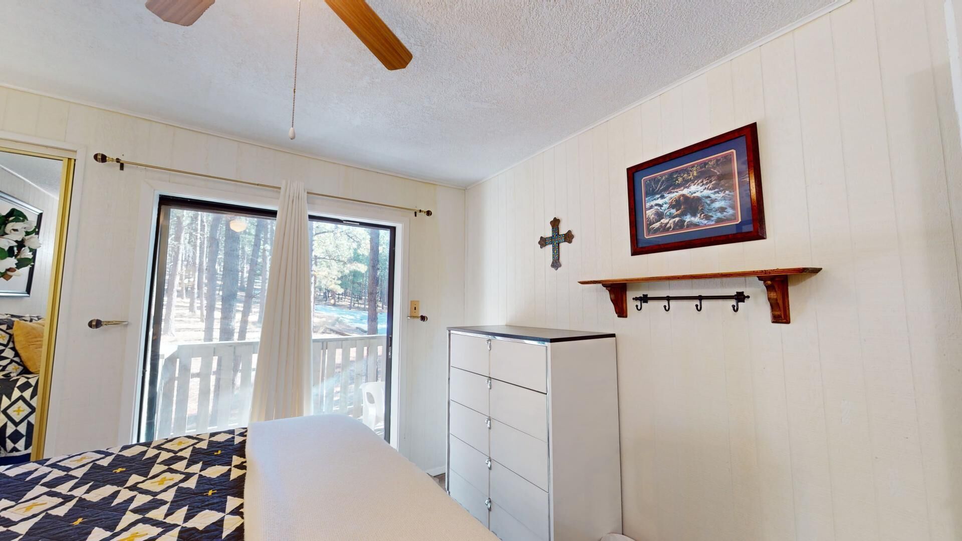 Une chambre à coucher comprenant un lit, une commode, une fenêtre avec des rideaux, un ventilateur de plafond et un tableau encadré accroché au mur.