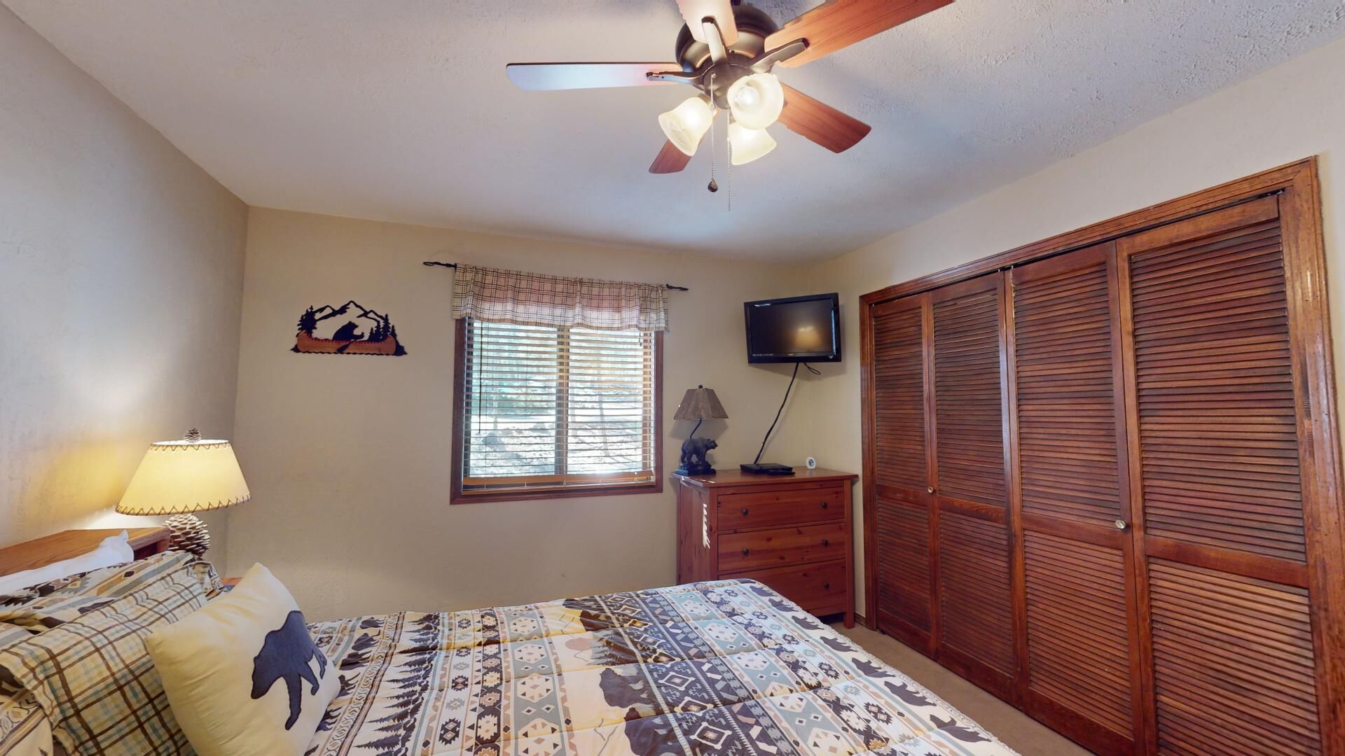 Ein Schlafzimmer mit Bett, Deckenventilator, Fernseher, Kleiderschrank und einem Fenster mit Jalousien.