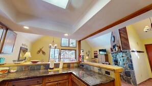 A spacious kitchen with granite countertops, wooden cabinets, and a stone fireplace. - Large Deck | Fireplace | Forest Views | Rose's Haven (Angel Fire)