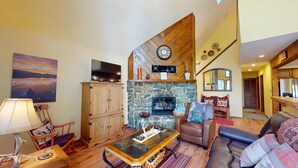 A cozy living room with a stone fireplace, wooden furniture, and a television on a cabinet. - Large Deck | Fireplace | Forest Views | Rose's Haven (Angel Fire)