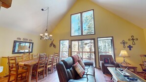 A spacious living room with a high ceiling, large windows, a dining area with wooden furniture, and a cozy seating area with a leather couch and chairs. - Large Deck | Fireplace | Forest Views | Rose's Haven (Angel Fire)
