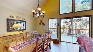 A dining area with a wooden table, chairs, and a chandelier. There is a large window with a view of trees. - Large Deck | Fireplace | Forest Views | Rose's Haven (Angel Fire)