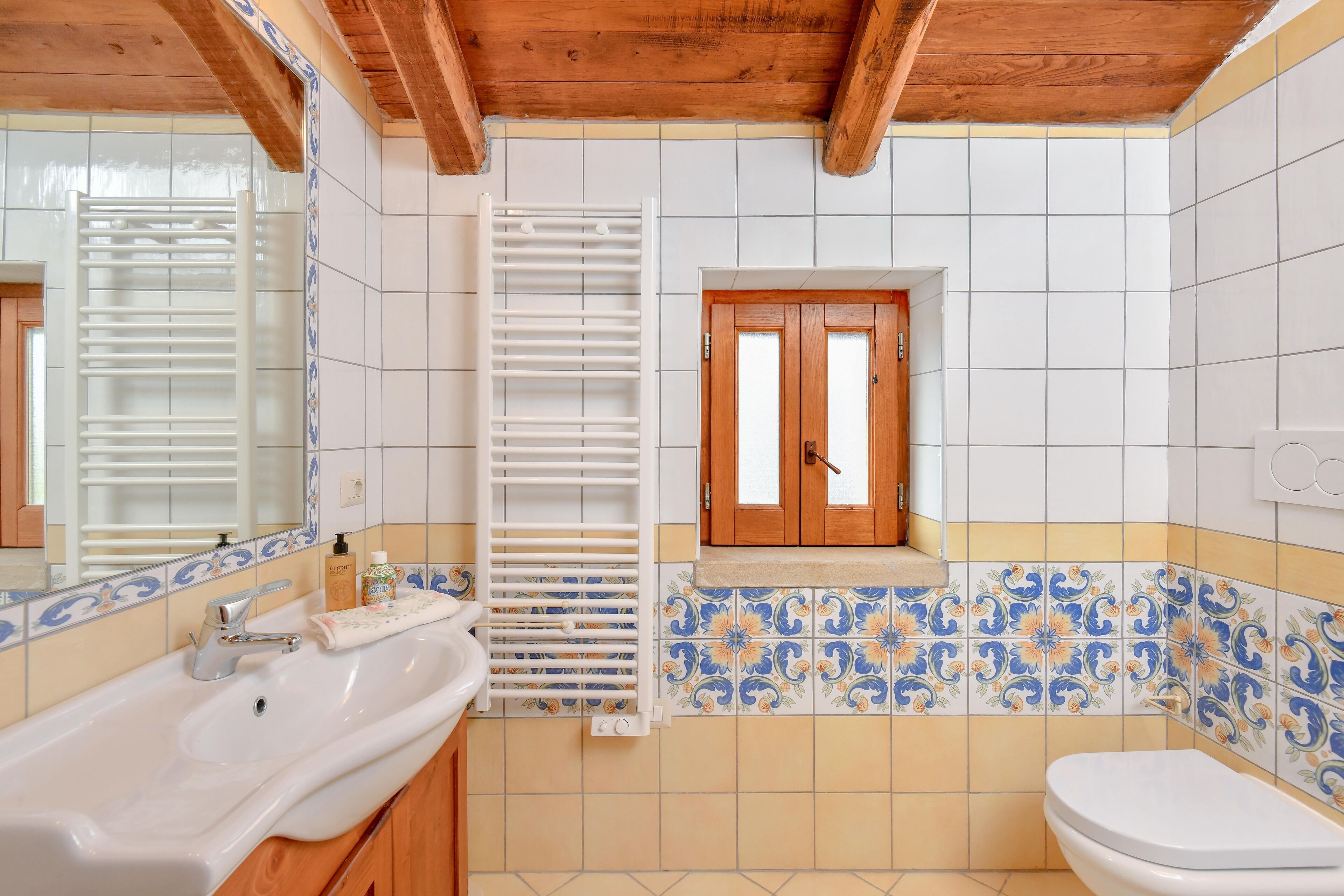 A bathroom with a white sink, wooden cabinets, a window with wooden shutters, and tiled walls with a floral pattern.