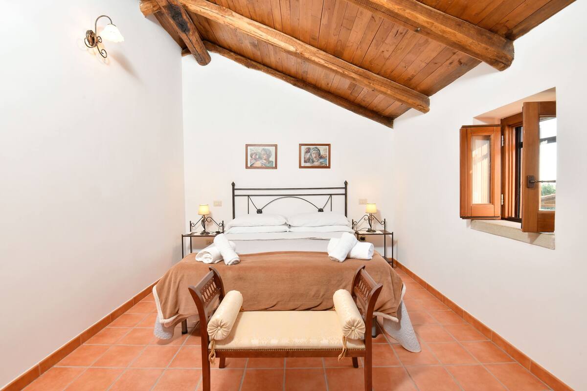 A bedroom with a wooden ceiling, a bed with a headboard, two bedside tables with lamps, and a window with a view.