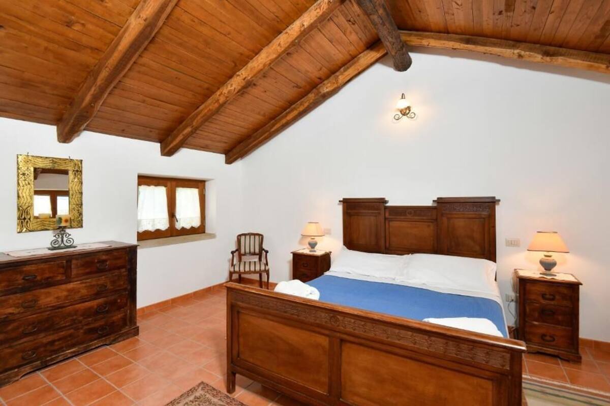 A bedroom with a wooden ceiling, a bed with a blue bedspread, two bedside tables with lamps, a wooden dresser, a mirror with a decorative frame, and a window with curtains.