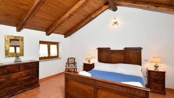 Une chambre avec un plafond en bois, un lit recouvert d’une courtepointe bleue, deux tables de chevet avec des lampes, une commode en bois, un miroir avec un cadre décoratif et une fenêtre avec des rideaux.