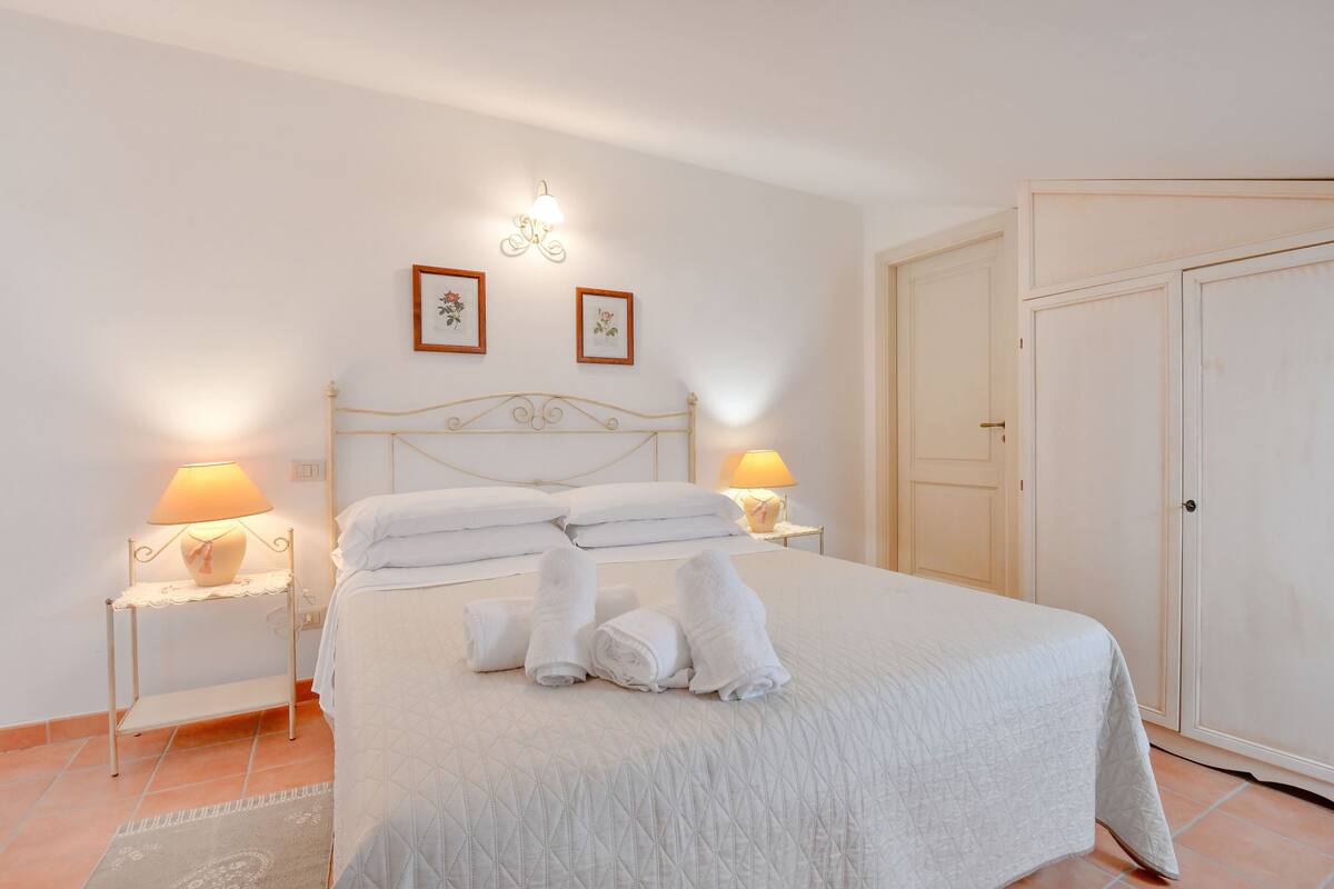 A neatly made bed with white linens and towels, a bedside table with a lamp, and a wall-mounted light fixture.