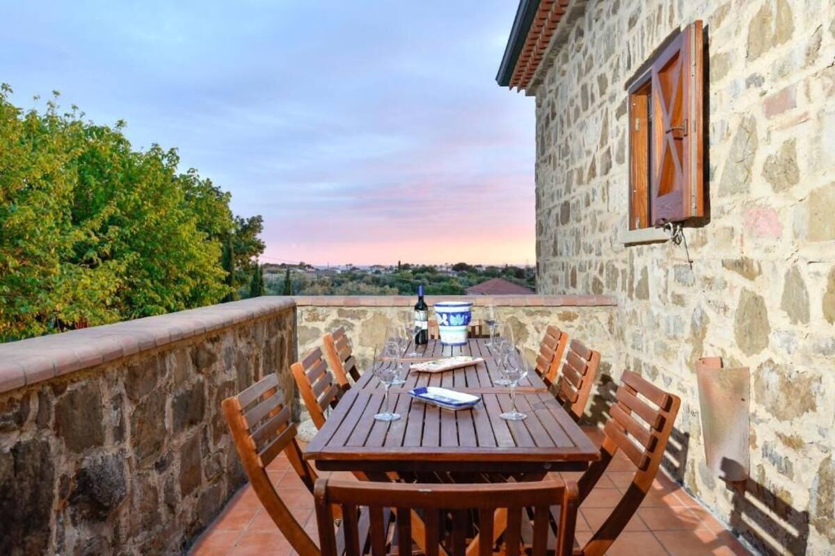 A stone building with a wooden table and chairs, a window, and a view of greenery.
