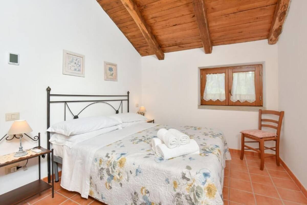 A bedroom with a wooden ceiling, a bed with a floral bedspread, a chair, and a window with curtains.