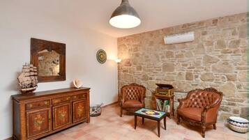 A living room with a stone wall, a wooden cabinet with floral designs, two leather armchairs, a small table with a colorful tabletop, and a hanging pendant light.