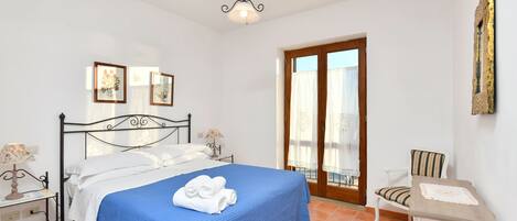A bedroom with a bed, a chair, a small table, a ceiling light, and a door leading to a balcony.