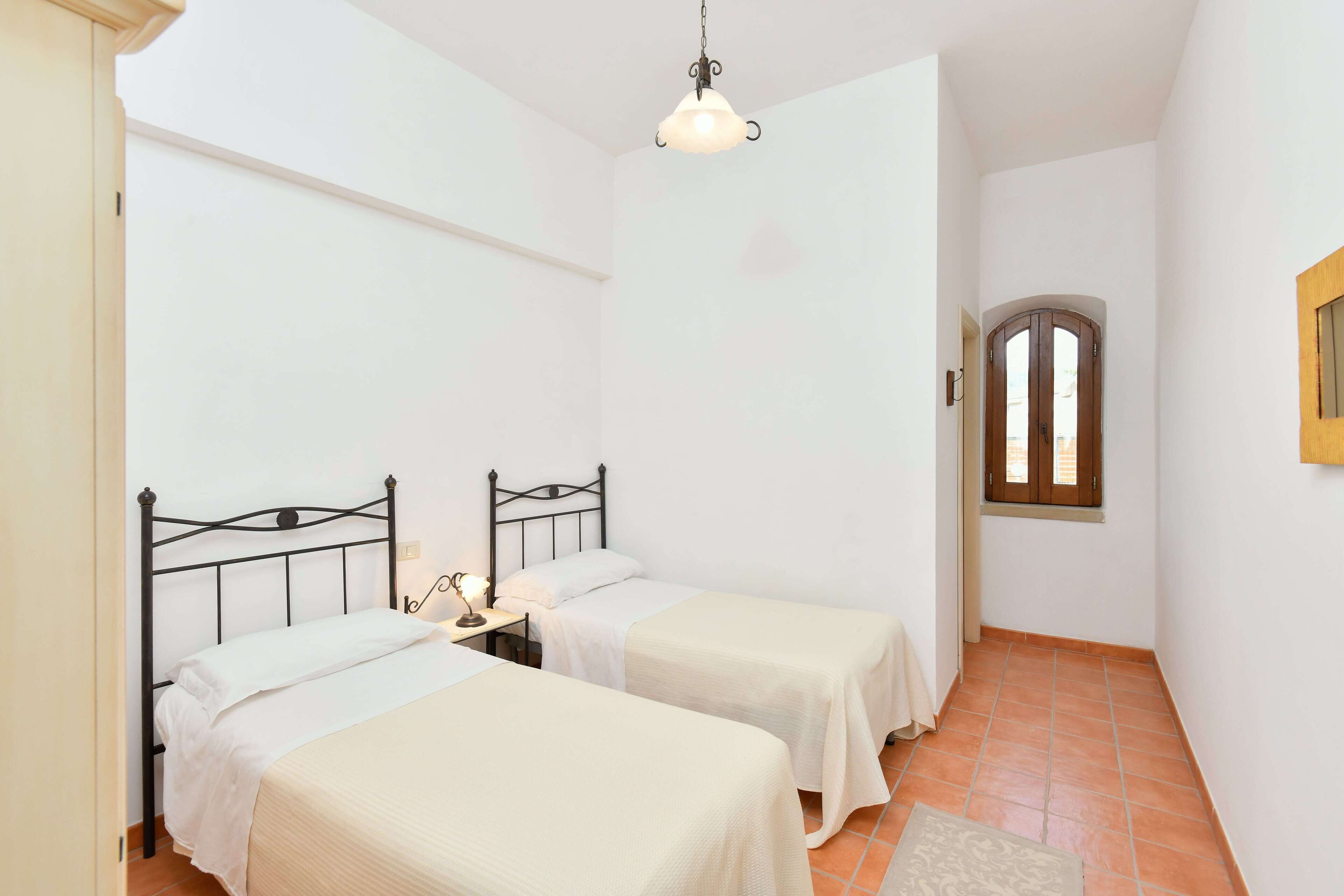 A small bedroom with two single beds, a nightstand, a hanging light fixture, and a window with a wooden frame.