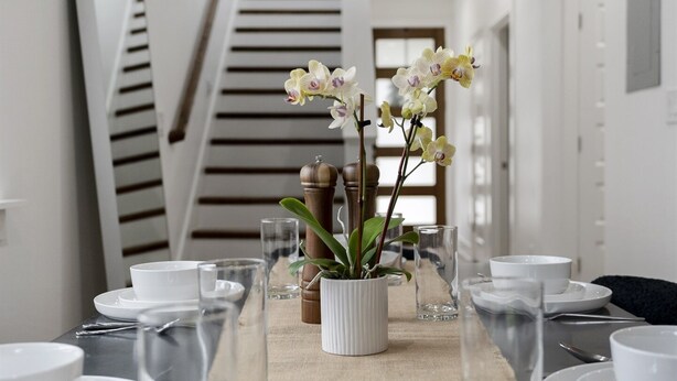 Une table à manger dressée avec de l’assiette, des bols et des verres blancs, et une orchidée en pot placée au centre.