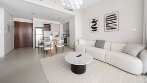 A modern living room with a white sofa, a round coffee table, and a dining area with chairs and a table. - Stylish 1 BD Near the Lagoon in Creek Harbour (Dubai)