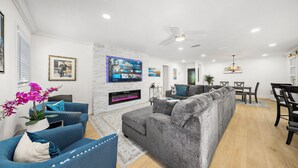 A modern living room with a grey sectional sofa, blue armchairs, a flat-screen TV mounted on the wall, a fireplace, and a dining area with a table and chairs. - Ultimate Vacation Spot: Pool, Mini Golf & Hammock! (Tampa)