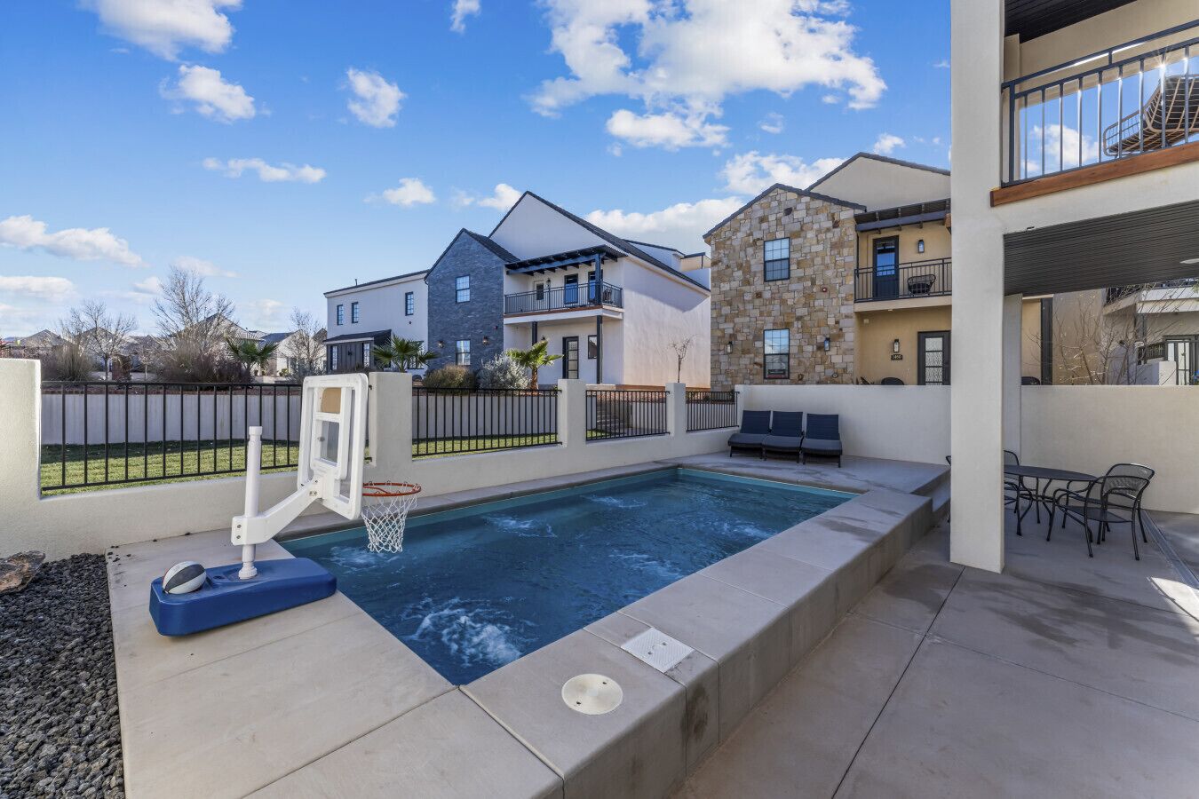 A swimming pool with a basketball hoop, a patio area with a table and chairs, and a view of neighboring houses.
