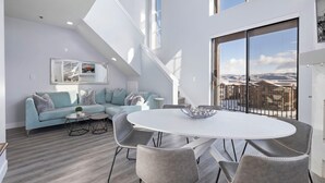 Una sala moderna con un sofá azul claro, sillas grises, una mesa de comedor blanca y un amplio ventanal con vistas a las montañas.