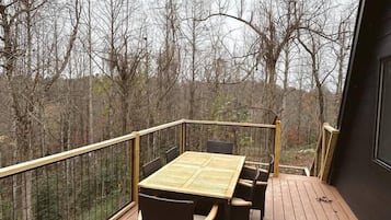 Een houten terras met een tafel en stoelen, omringd door een metalen reling, met uitzicht op een bebost gebied.