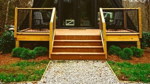 A modern, dark-colored cabin with a triangular roof, a wooden deck with a glass railing, and a number "620" above the entrance.