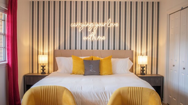 A hotel room with a striped wallpaper, a bed with white and yellow bedding, two bedside lamps, and a neon sign above the bed. - River Run Retreat - Cowboy Pool, Fire Pit & Pool Table! Close to Campus & River (San Marcos)