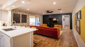 A modern kitchen with a white countertop and sink, a dining area with a red sofa and yellow chairs, and a hallway with wooden flooring and a ceiling fan. - River Run Retreat - Cowboy Pool, Fire Pit & Pool Table! Close to Campus & River (San Marcos)