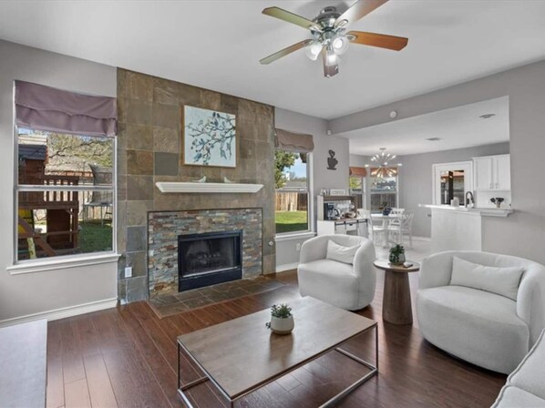 open-concept living space features comfortable white armchairs, a classic stone fireplace, and a seamless view right into the kitchen and dining area.