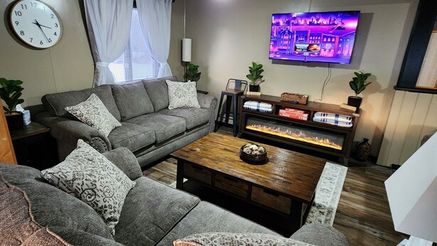 Una sala de estar con un sofá gris, una mesa de centro de madera, un televisor de pantalla plana, una chimenea y un reloj en la pared.