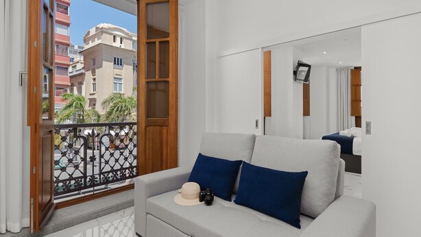 Una sala moderna con un sofá gris, un sombrero y una cámara encima. Hay una puerta de madera con paneles de vidrio y vistas a edificios.