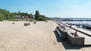 A sandy beach with a wooden pier extending into calm waters, surrounded by greenery and houses in the background. - Strandläufer by Interhome (Seebad Altefähr)