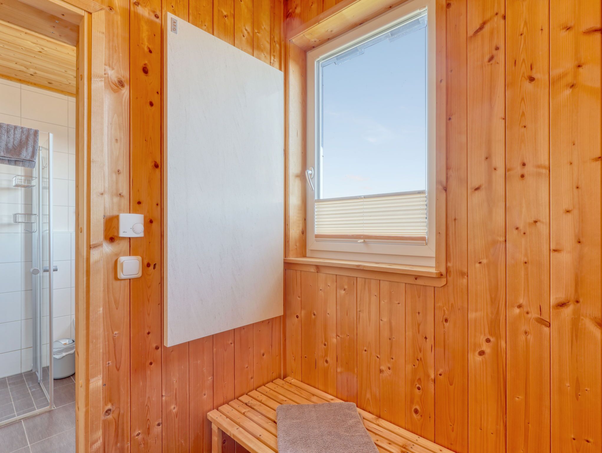 A wooden sauna room with a bench, a window with blinds, and a thermostat on the wall.