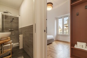 A modern bathroom with a shower, sink, and toilet. There is a bed in the adjacent room with a view of buildings through a window.
