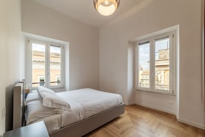 A bedroom with a bed, two windows, a wooden floor, and a ceiling light.
