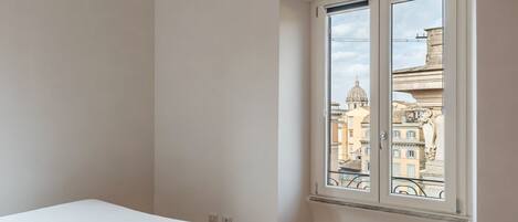 A bedroom with a bed, a large window with a view of buildings, and a wooden floor.
