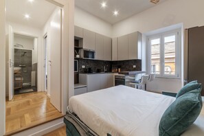 A modern bedroom with a large bed, a small desk, and a kitchenette.