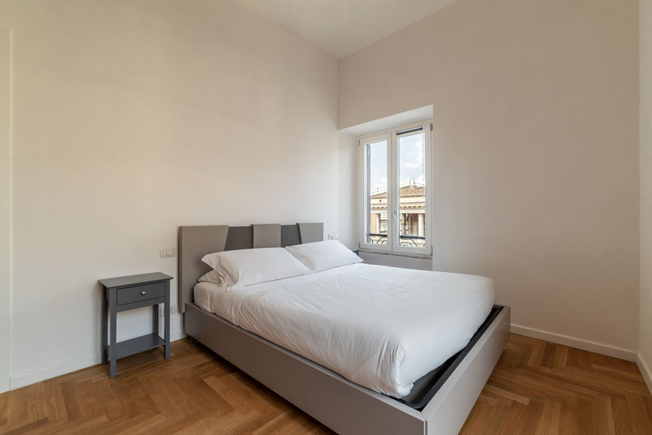 A bedroom with a bed, a nightstand, and a window with a view of buildings.