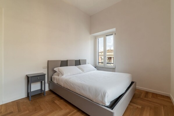 A bedroom with a bed, a nightstand, and a window with a view of buildings.