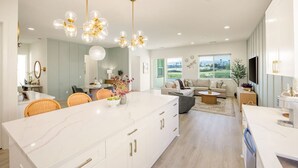 A modern kitchen with a central island, dining area with wicker chairs, and a living room with a sofa and coffee table. - Fairway Gem: Pool+Spa, Putting Green, EV Charger! (Indio)