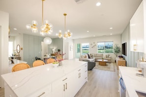 A modern kitchen with a central island, dining area with wicker chairs, and a living room with a sofa and coffee table. - Fairway Gem: Pool+Spa, Putting Green, EV Charger! (Indio)