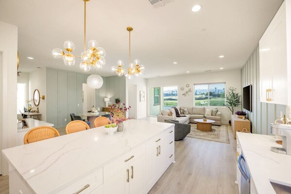 A modern kitchen with a central island, dining area with wicker chairs, and a living room with a sofa and coffee table.