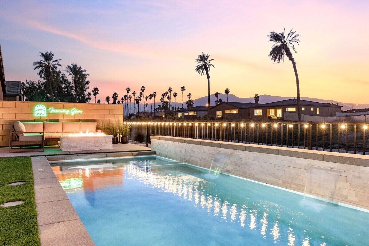 A swimming pool with a waterfall feature, surrounded by a brick wall and palm trees, with a neon sign in the background.