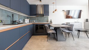 Une cuisine moderne avec des armoires bleu foncé, une hotte de cuisinière en acier inoxydable et un four encastré. On y trouve également un coin repas avec une table et des chaises, ainsi qu’un téléviseur à écran plat fixé au mur.