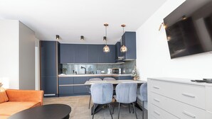 Une cuisine moderne avec des meubles de couleur bleu foncé, un coin repas avec une table et des chaises, un canapé gris et un téléviseur fixé au mur.