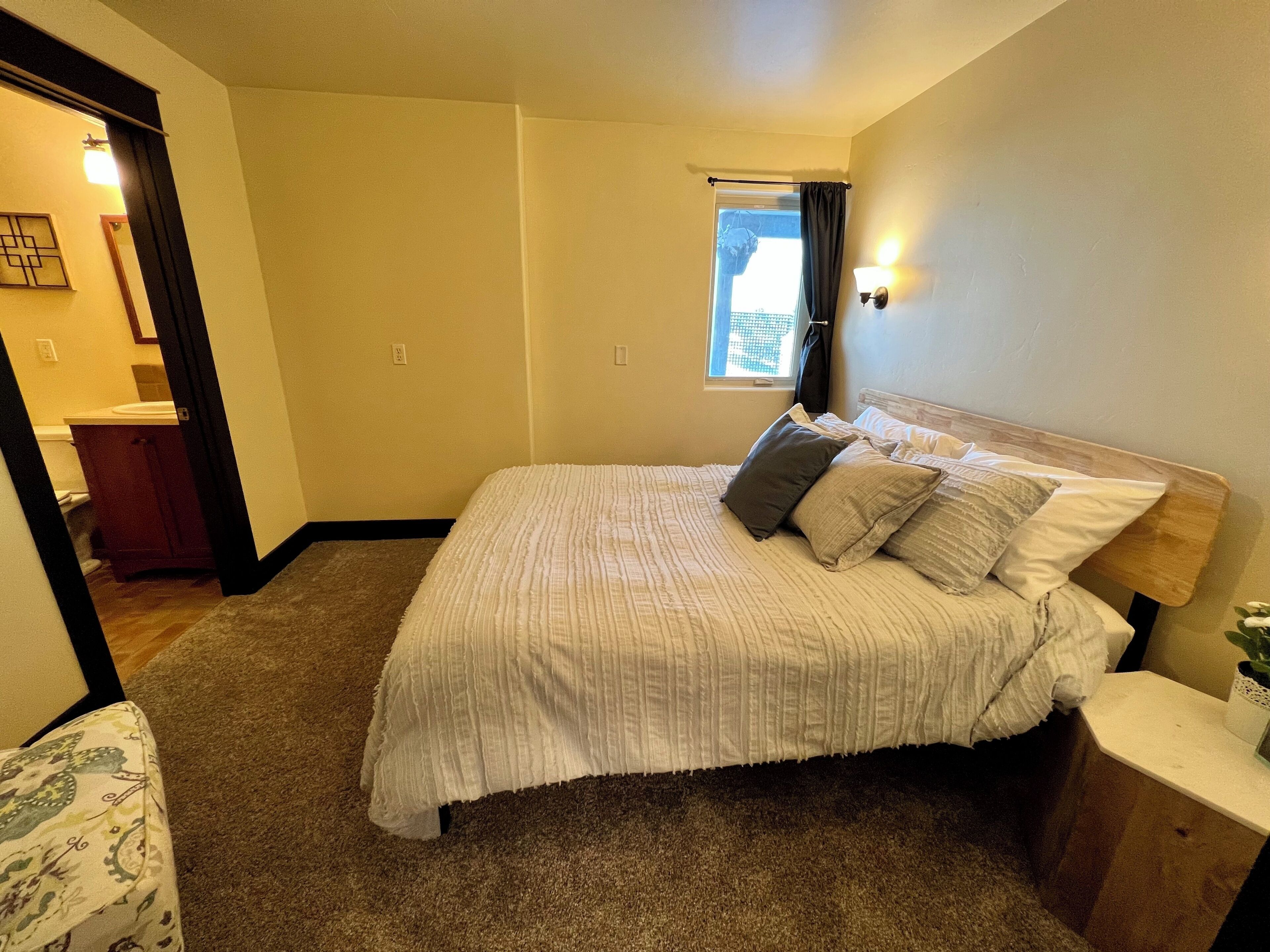 Quarto com cama, mesa de cabeceira, janela e porta para um banheiro.