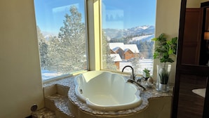 Ett badrum med ett fristående badkar, ett fönster med utsikt över snötäckta hus och träd, och en liten växt på fönsterbrädan.
