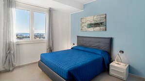 A bedroom with a bed, a nightstand, a wall-mounted lamp, a window with curtains, and a painting. - Apartment 'Iris' with Sea View, Private Terrace and Air Conditioning (Albenga)