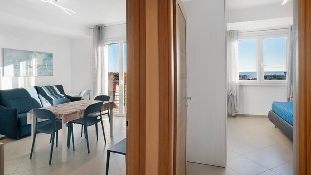 A modern living room with a sofa, dining table, and chairs, a balcony with a view, and a door leading to another room. - Apartment 'Iris' with Sea View, Private Terrace and Air Conditioning (Albenga)