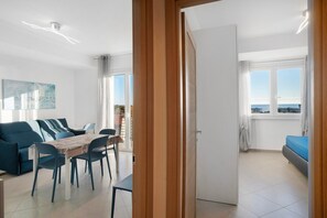 A modern living room with a sofa, dining table, and chairs, a balcony with a view, and a door leading to another room.