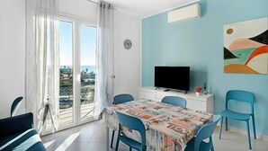 A dining area with a table, chairs, a TV, and a view through the window. - Apartment 'Iris' with Sea View, Private Terrace and Air Conditioning (Albenga)